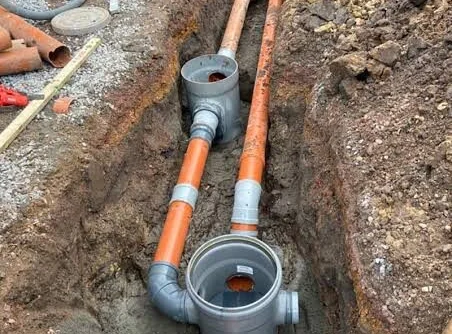 Twee inspectieputten en oranje PVC-rioolbuizen in een uitgegraven sleuf.