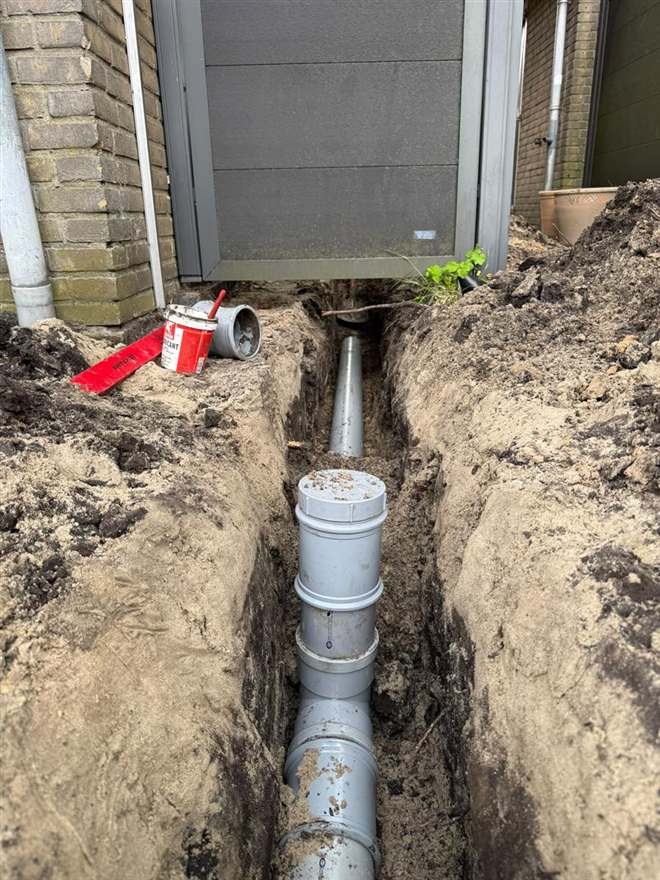 Uitgegraven sleuf langs een woning met een nieuw aangelegde grijze PVC-rioolleiding en een inspectieput.